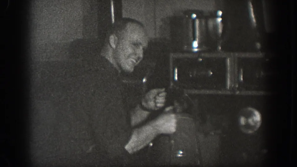 A man pauses to smile while working near a large dark appliance in a home movie from Chelan, Washington, dated 1939.