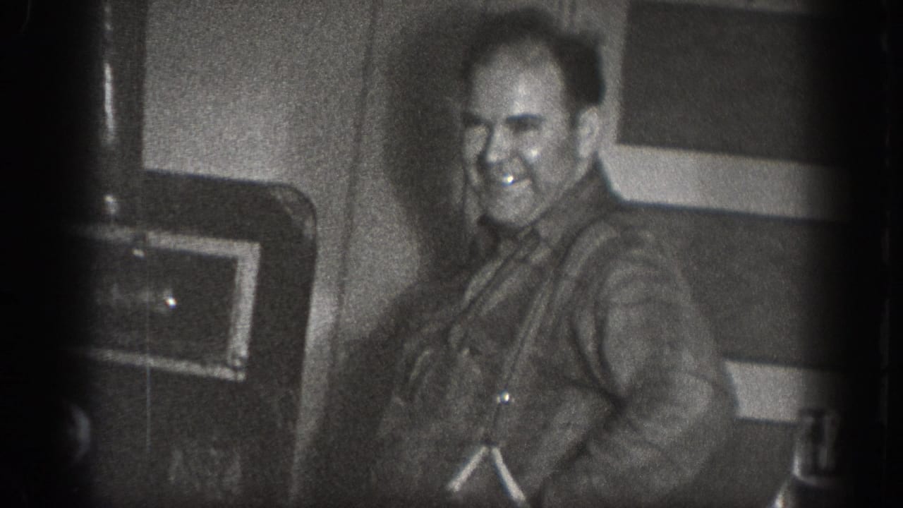A man smiles directly at the camera inside a private space with dark mechanical equipment behind him; from a 1939 home movie from Chelan, Washington.