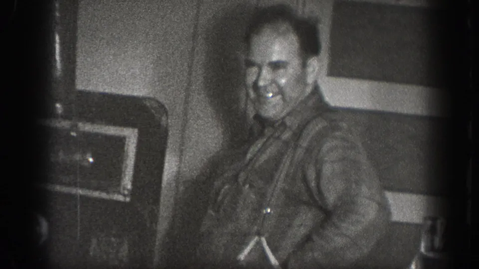 A man smiles directly at the camera inside a private space with dark mechanical equipment behind him; from a 1939 home movie from Chelan, Washington.