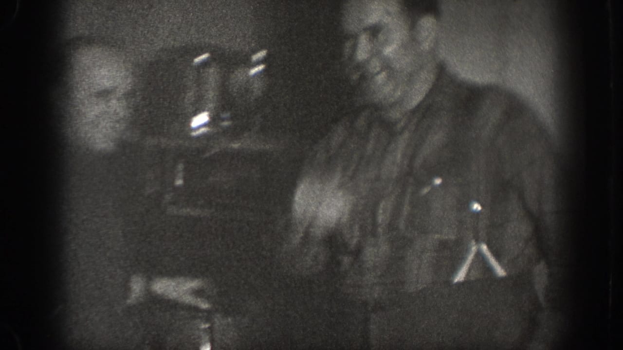 Two men in a dimly lit interior; one smiling, metallic objects visible at left in background.