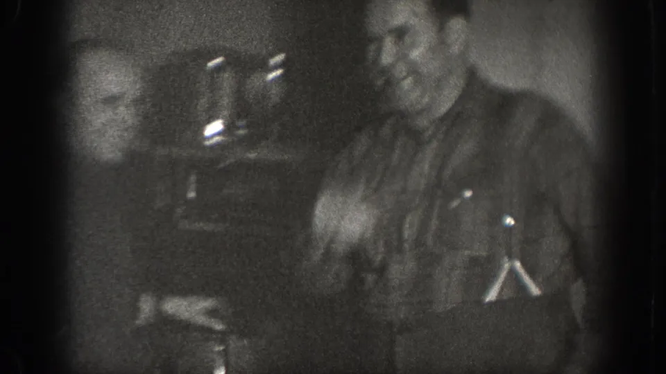 Two men conversing in a dark interior, one smiling; a metallic object at left may show the stove referenced in the title, 1939 8mm home movie, Chelan WA.