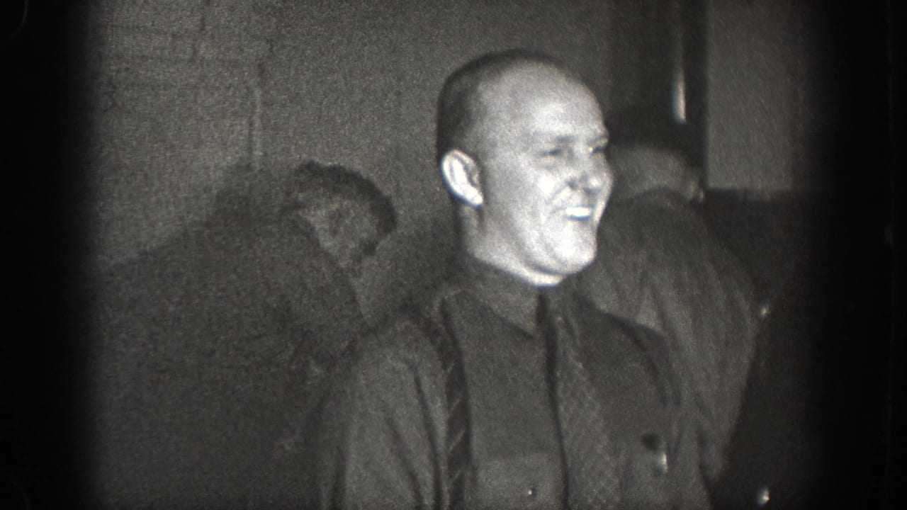 Bald man in dark button-up shirt laughing broadly, second man partially visible behind him; dimly lit private interior, 1930s 8mm home movie, Chelan WA.