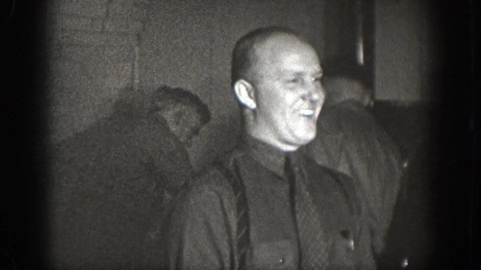 Bald man in dark button-up shirt laughing broadly, second man partially visible behind him; dimly lit private interior, 1930s 8mm home movie, Chelan WA.