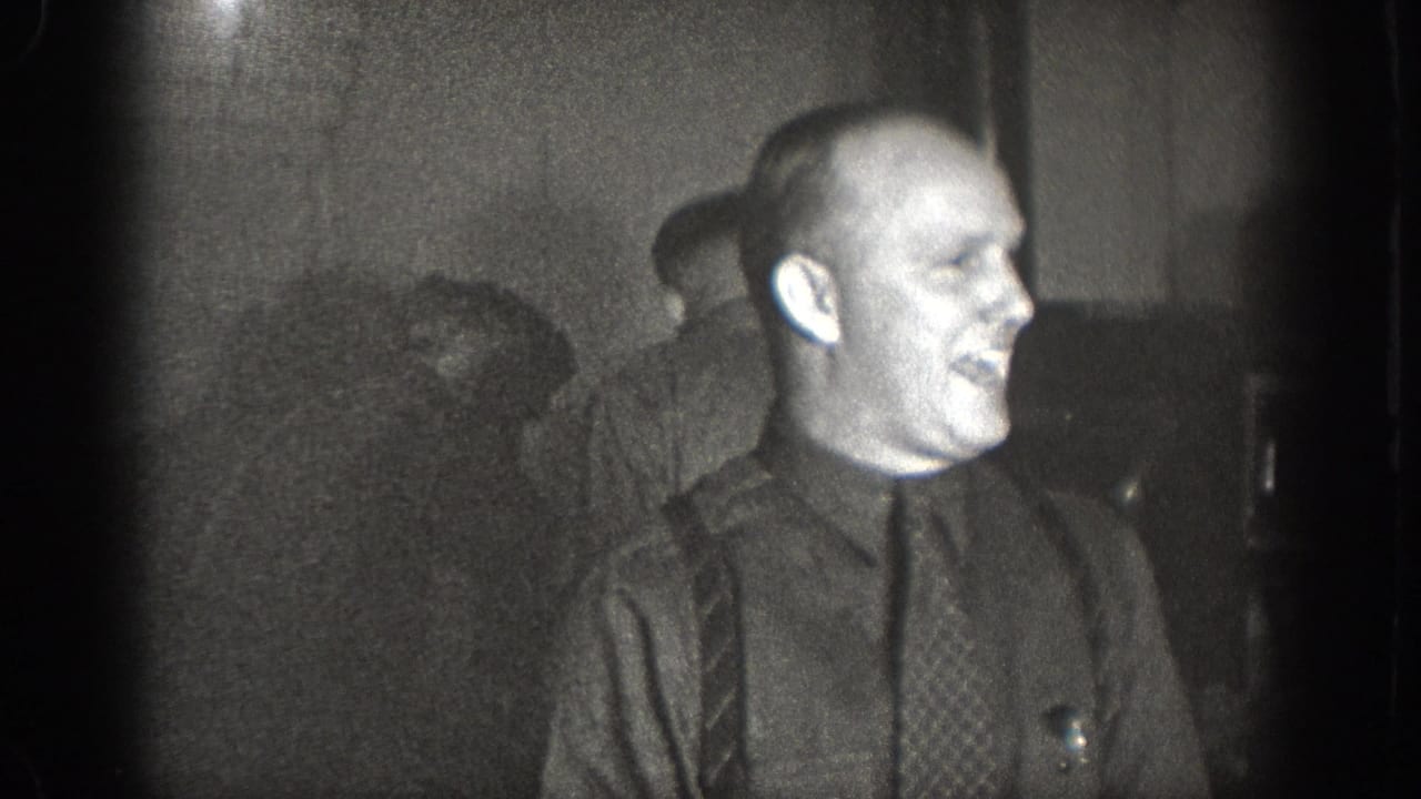 Bald man in profile wearing dark shirt, patterned necktie, and suspenders, laughing; second man partially visible at left; 1930s 8mm home movie, Chelan WA.