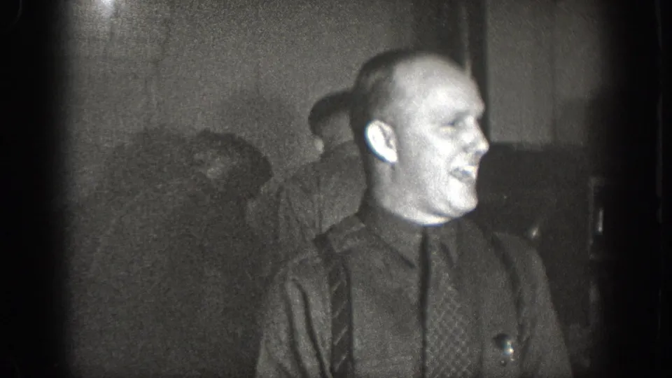 Bald man in profile wearing dark shirt, patterned necktie, and suspenders, laughing; second man partially visible at left; 1930s 8mm home movie, Chelan WA.