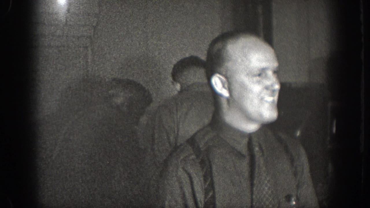 Bald man in dark shirt and patterned tie smiling, second figure visible behind him; dim private interior, 1930s 8mm home movie, Chelan WA.