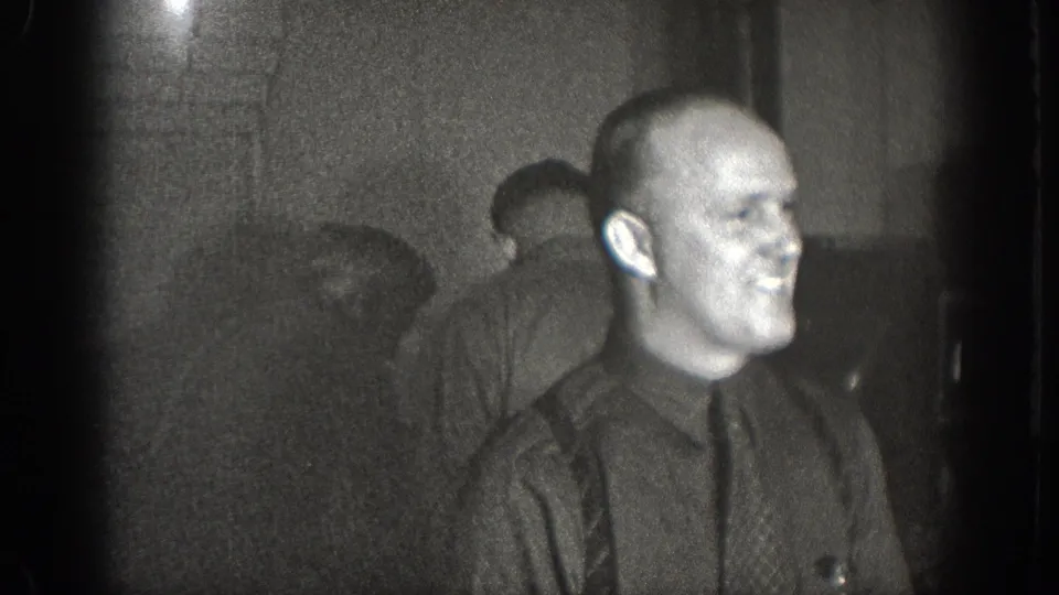 Bald man in dark shirt and patterned tie smiling, second figure visible behind him; dim private interior, 1930s 8mm home movie, Chelan WA.