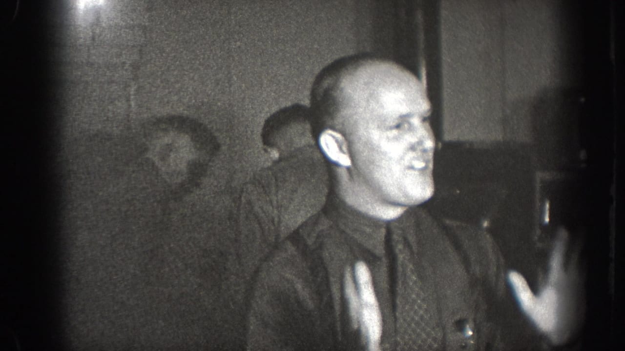 Bald man in dark shirt and necktie gesturing animatedly while seated at a table, interior of what may be a bar or diner, Chelan Washington circa 1939, 8mm film.