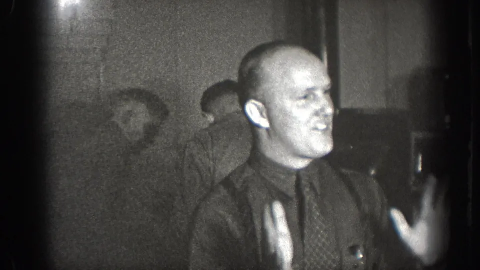 Bald man in dark shirt and necktie gesturing animatedly while seated at a table, interior of what may be a bar or diner, Chelan Washington circa 1939, 8mm film.