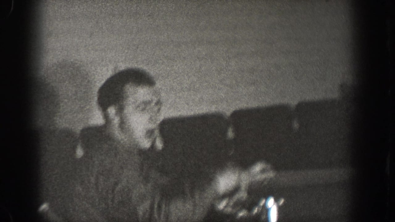 Man seated at table with hand resting near a chrome vessel, booth seating visible behind, black-and-white 8mm interior.