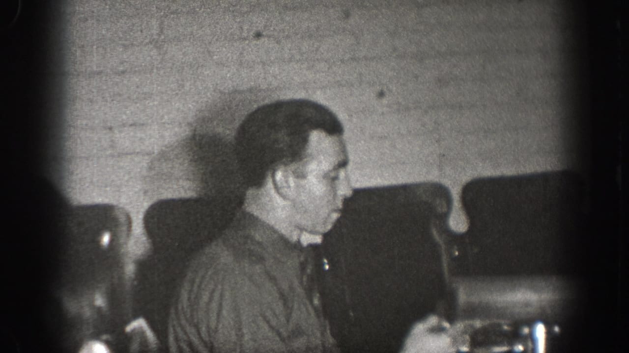 Adult male in dark work shirt, seated in profile at a table in a brick-walled interior, examining a small device; late-1930s home movie, Chelan, Washington.