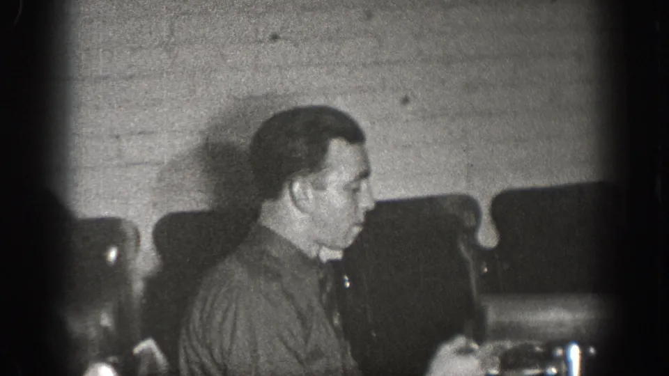 Adult male in dark work shirt, seated in profile at a table in a brick-walled interior, examining a small device; late-1930s home movie, Chelan, Washington.