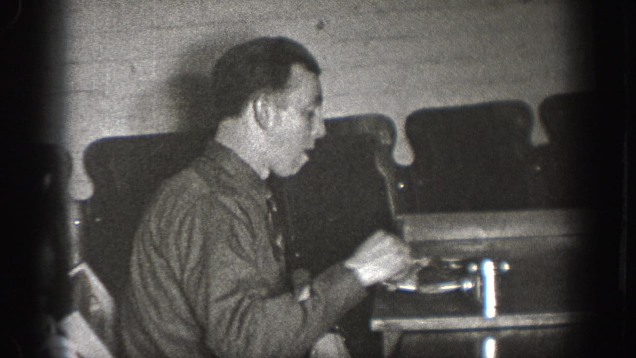 Man in dark work shirt, face partially visible, hands near a small object on the table; several upholstered chairs in background.