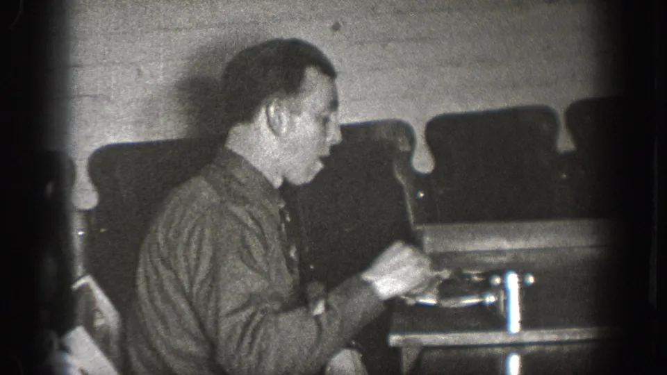 Adult male, face partially toward camera, hands raised near a small device on a table in a brick-walled interior; late-1930s home movie footage, Chelan, Washington.