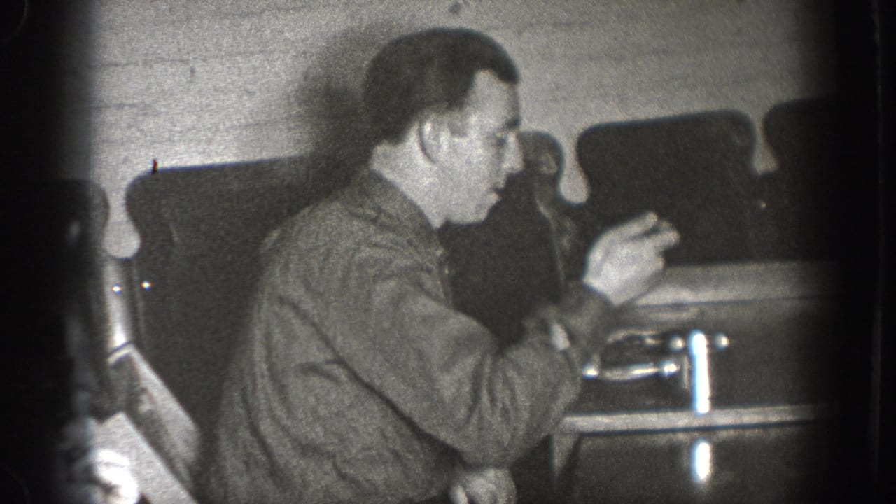 Adult male in close profile with both hands raised above table surface, handling a small mechanical object; interior with brick wall and dark chairs, consistent with late-1930s home movie.