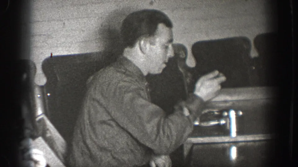 Adult male in close profile with both hands raised above table surface, handling a small mechanical object; interior with brick wall and dark chairs, consistent with late-1930s home movie.