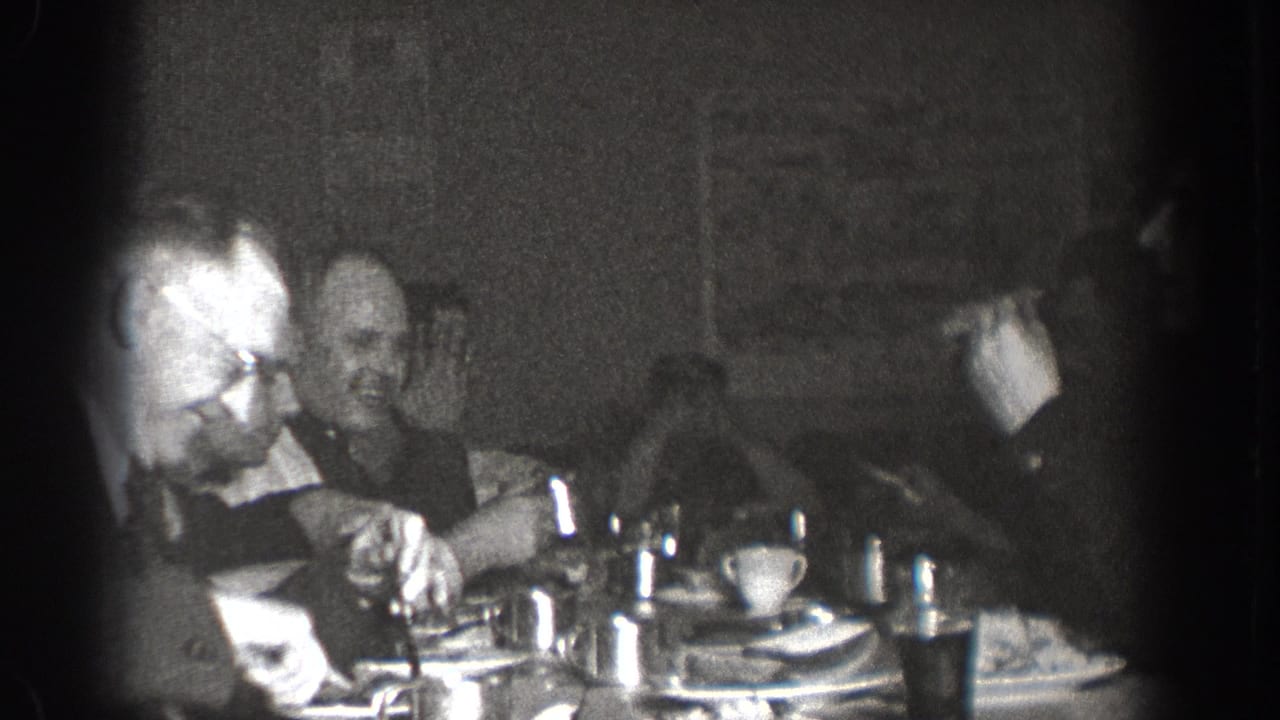 Group of men in suits at a communal dining table in Chelan, Washington, circa 1939; cups, plates, and glassware visible in grainy 8mm footage.
