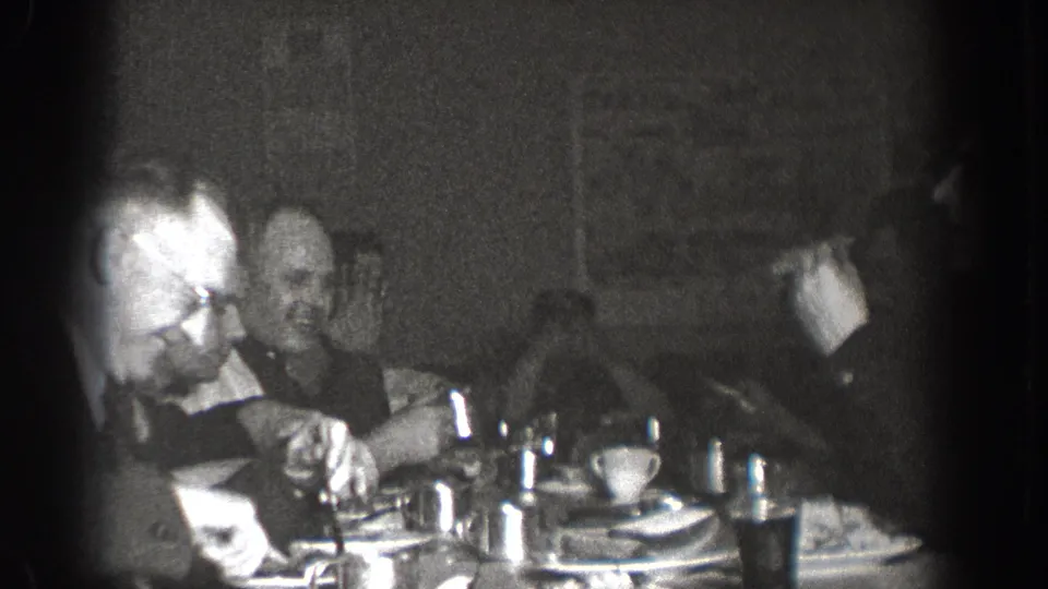 Group of men in suits at a communal dining table in Chelan, Washington, circa 1939; cups, plates, and glassware visible in grainy 8mm footage.