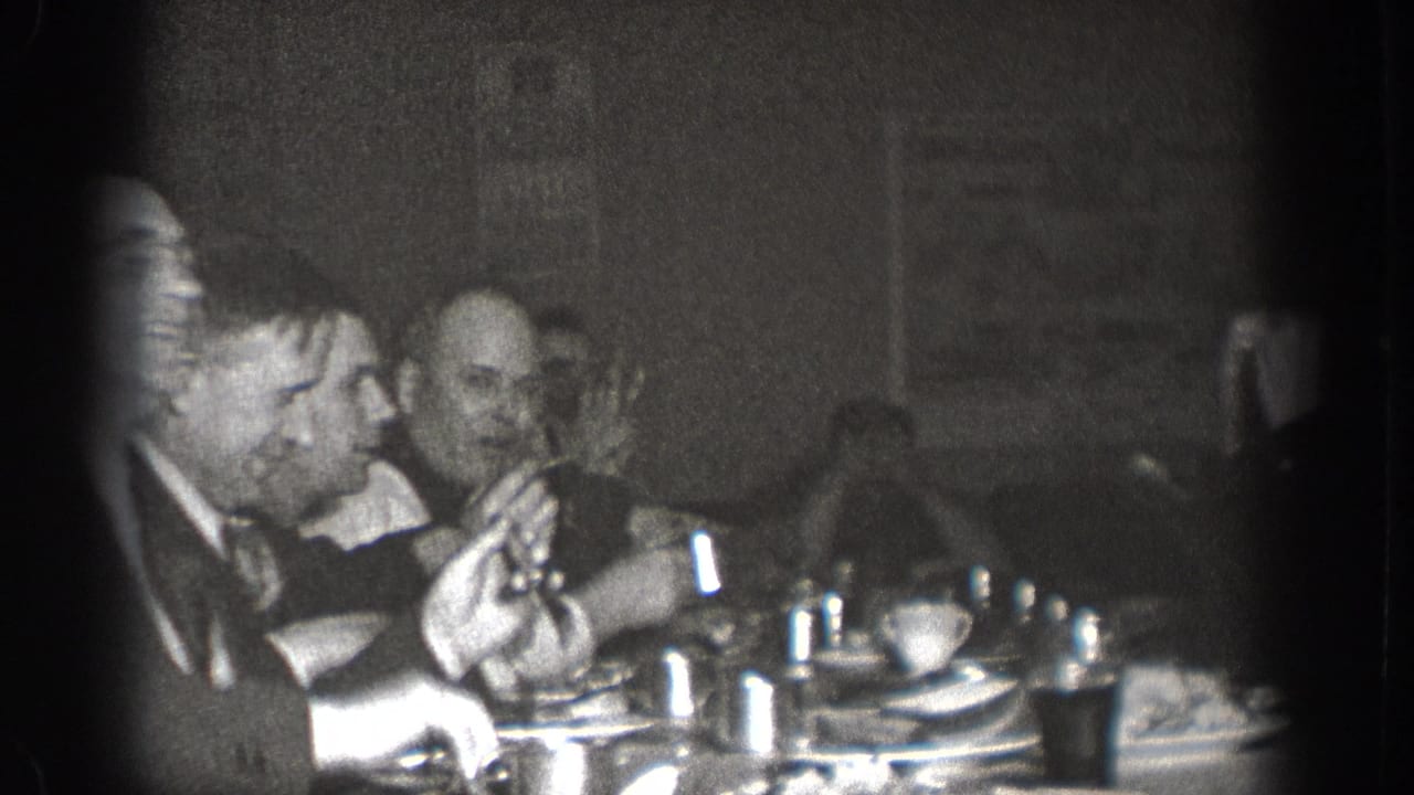 Two men in dark suits at a dining table; one raises a hand in conversation; cups and plates on table.