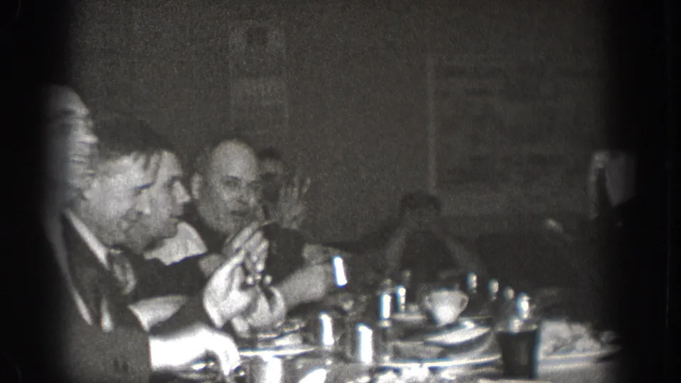 Two men in dark suits at a communal dinner table in Chelan, Washington, circa 1939; one raises a hand mid-conversation.
