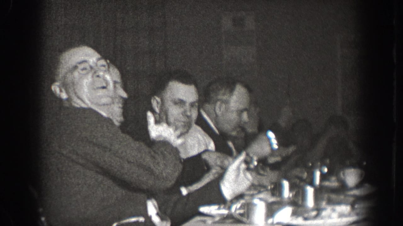 Several men in suits seated at a dining table, one laughing broadly, cups and tableware visible, black and white 8mm film.