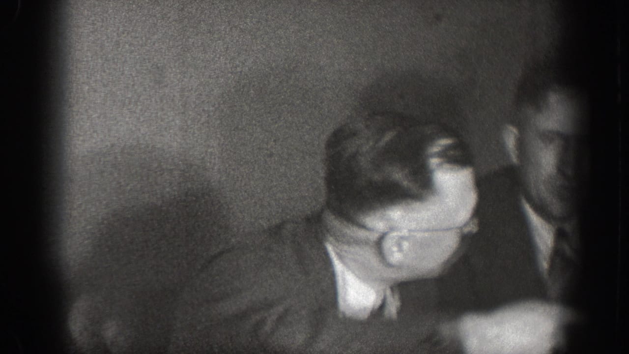 Dark, heavily vignetted frame of men at a dining table — underexposure and grain typical of 8mm home movie film stock, circa 1939 Chelan, Washington.