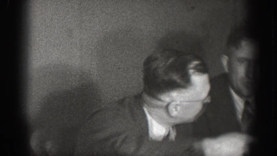 Dark, heavily vignetted frame of men at a dining table — underexposure and grain typical of 8mm home movie film stock, circa 1939 Chelan, Washington.