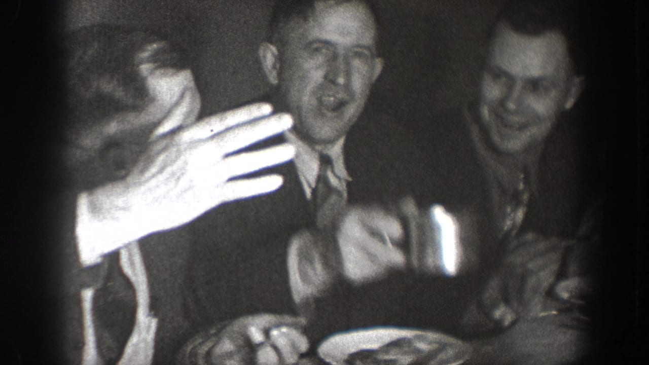 Animated group of suited men at a dining table, one reaching toward the camera with plates visible — candid 8mm home movie circa 1939, Chelan, Washington.