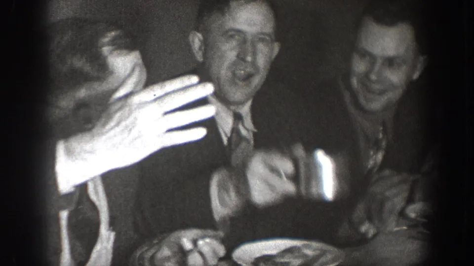 Animated group of suited men at a dining table, one reaching toward the camera with plates visible — candid 8mm home movie circa 1939, Chelan, Washington.
