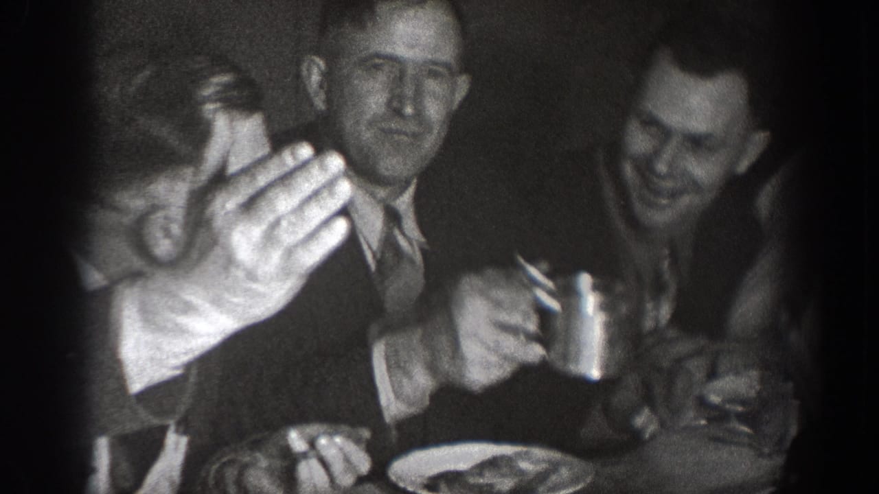 Two men at a table smiling, one raising a cup toward the camera in a toast, dinner plates visible, black and white 8mm.