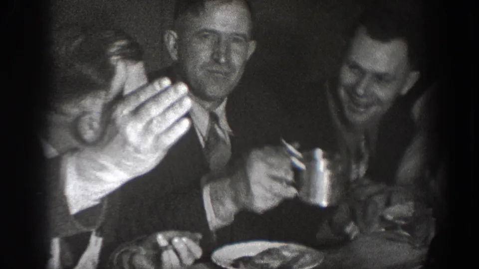 Two men laughing and raising cups in a toast at a dimly lit table with plates of food; candid 8mm home-movie moment consistent with 1930s Chelan, Washington.