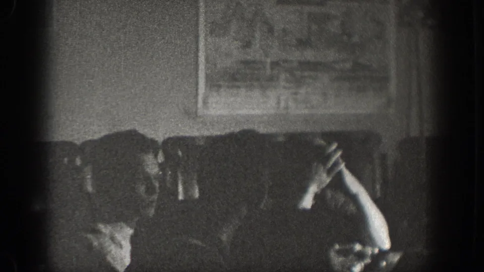 Dimly lit interior with seated figures at a table and what appears to be a framed poster or sign on the wall; heavily underexposed 8mm home-movie frame from a bar or gathering space.