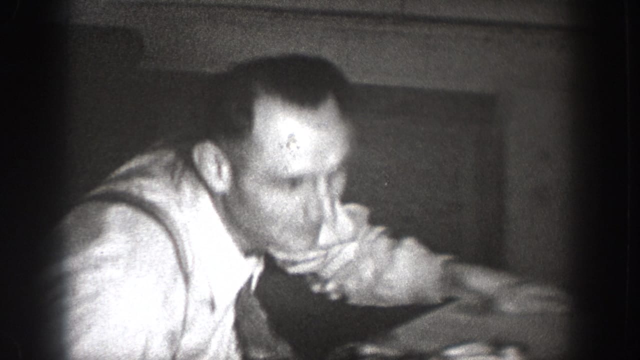 Close-up of a man in a light shirt leaning forward in a dark wood-paneled interior room, candid 8mm footage, Chelan, Washington, c.1939.