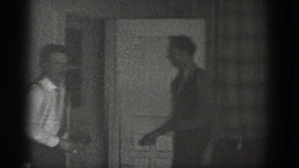 Two adult men standing in a dark interior near a doorway, one wearing a lighter shirt, candid 8mm footage from Chelan, Washington, c.1939.