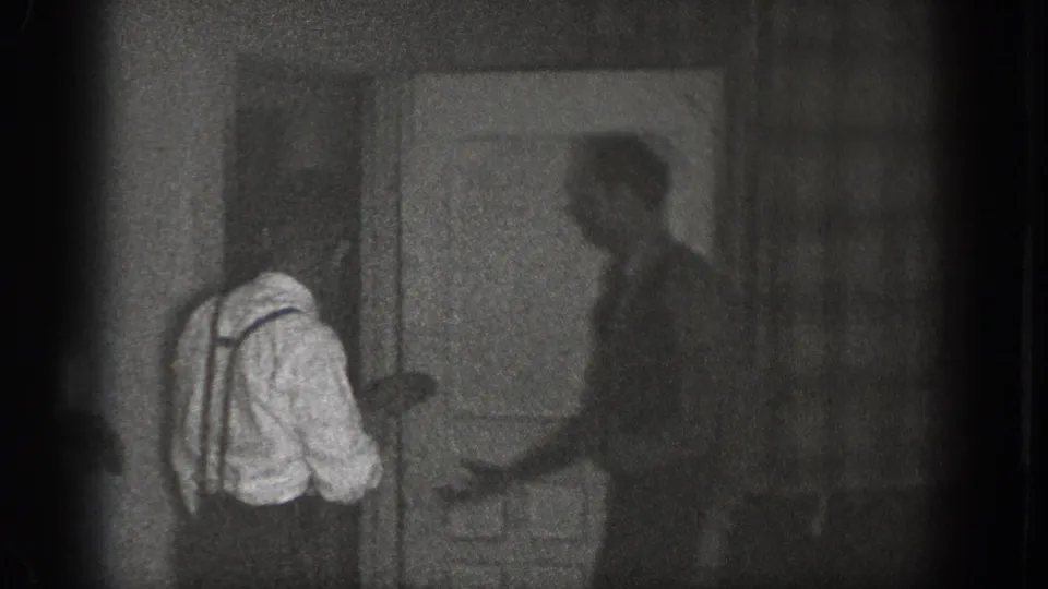 In this 1939 Chelan, Washington home-movie frame, several adult men stand in a dim interior near a doorway while a man in a white shirt and suspenders leans toward the group.