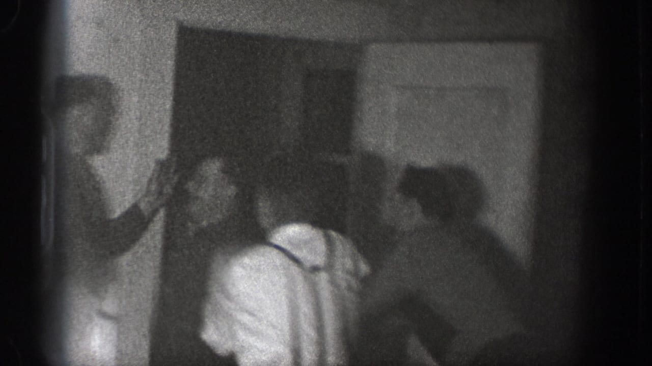 Several adult men appear clustered around a doorway in the Chelan home-movie scene, with one man in suspenders bent forward as others stand close by.