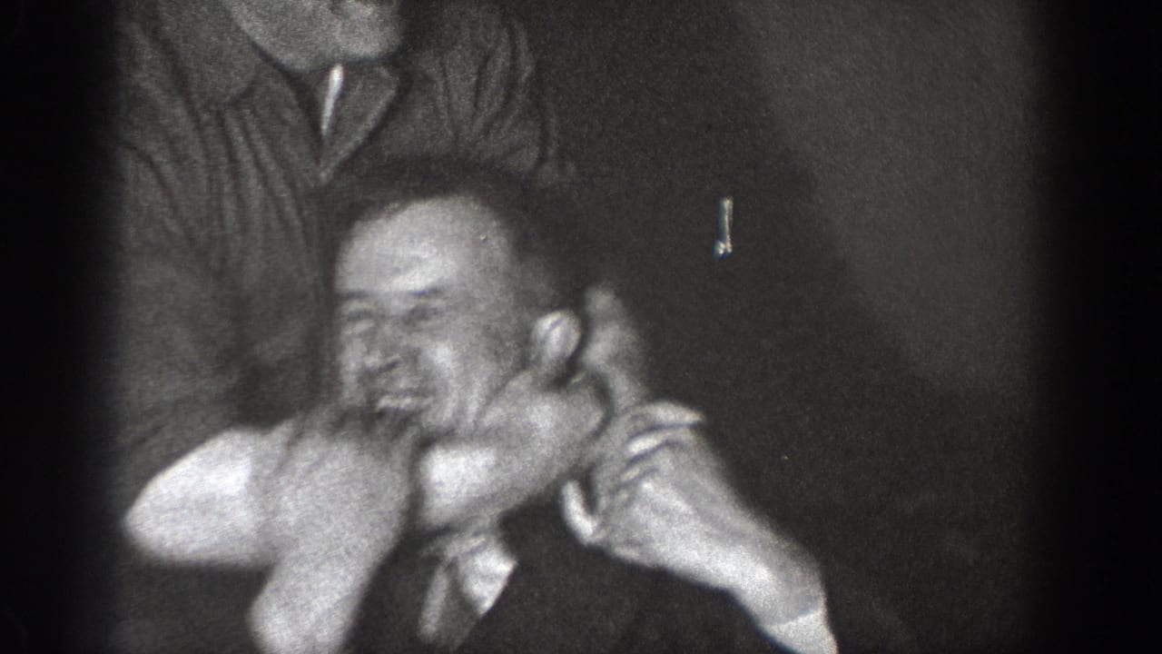 Two men in 1930s dress in a close-up playful face-grab, captured on 8mm home movie film, likely Chelan, Washington, circa 1939.