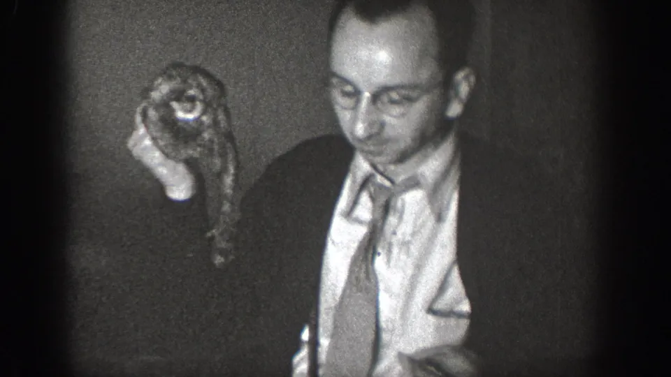 Man in suit and necktie holds a small live animal with large eyes toward the camera, in 8mm home movie footage, likely Chelan, Washington, circa 1939.