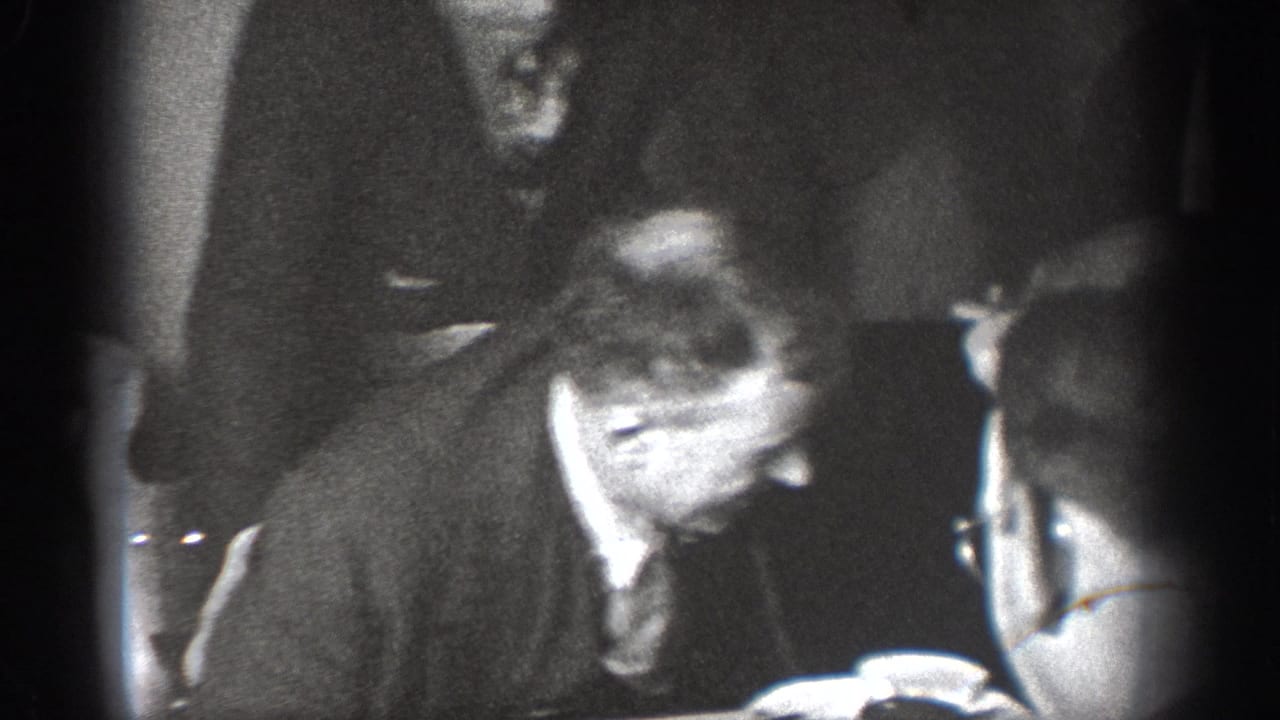 Two or three men indoors, one wearing glasses leaning over while another figure looks on, grainy 8mm black-and-white footage.