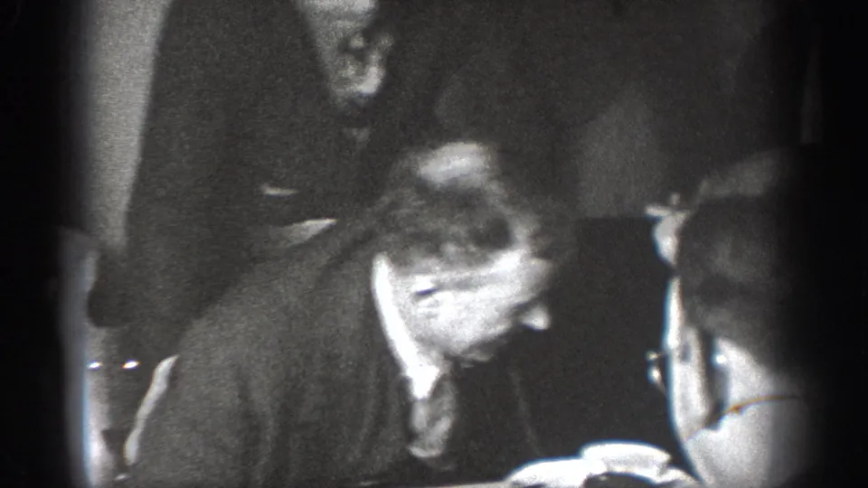 Men in dark suits gathered closely indoors, one with eyeglasses leaning forward, another figure watching; likely Chelan Washington circa 1939, 8mm home movie.