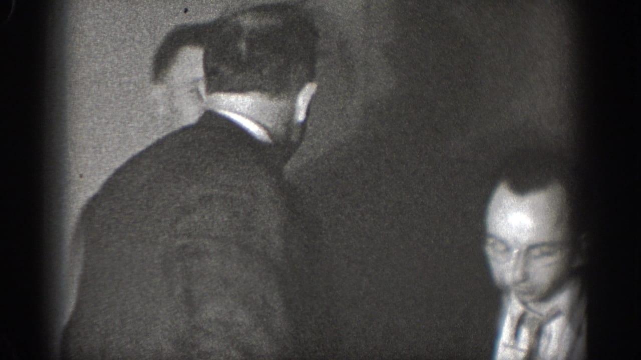 Two men in dark suits and ties in near-profile candid view indoors, likely Chelan Washington circa 1939; left-edge vignetting marks 8mm home movie format.