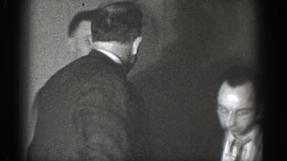Two men in dark suits and ties in near-profile candid view indoors, likely Chelan Washington circa 1939; left-edge vignetting marks 8mm home movie format.