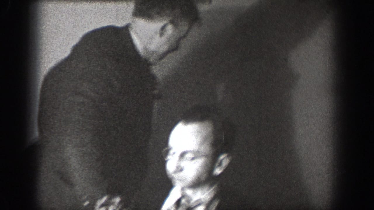 Man with eyeglasses and striped tie in candid indoor moment with a second figure leaning over from behind, likely Chelan Washington circa 1939, 8mm home movie.