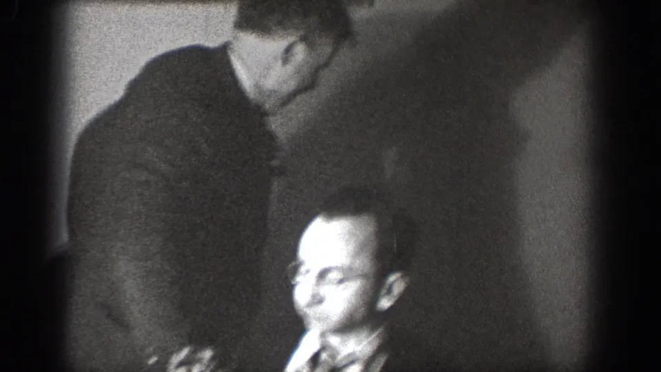 Man with eyeglasses and striped tie in candid indoor moment with a second figure leaning over from behind, likely Chelan Washington circa 1939, 8mm home movie.