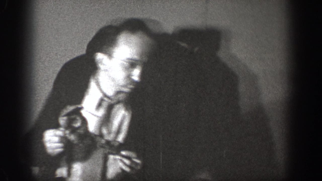 Amateur 8mm footage circa 1939, Chelan, Washington — man in dress shirt and tie examines a small dark object in a dimly lit indoor setting.
