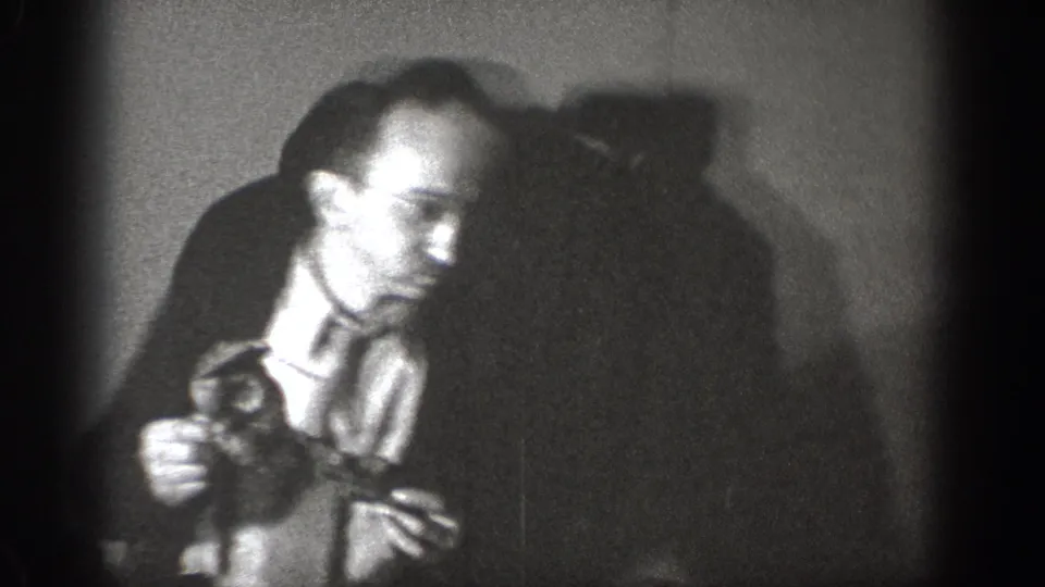 Amateur 8mm footage circa 1939, Chelan, Washington — man in dress shirt and tie examines a small dark object in a dimly lit indoor setting.
