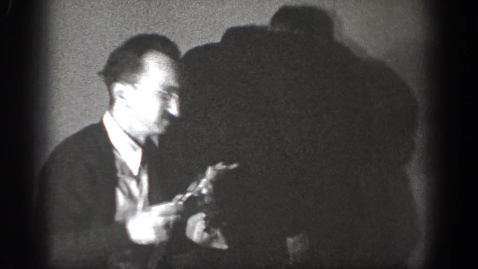 Amateur 8mm circa 1939, Chelan, Washington — bespectacled man in dark jacket holds a small dark object and smiles; second man partially visible in background shadow.