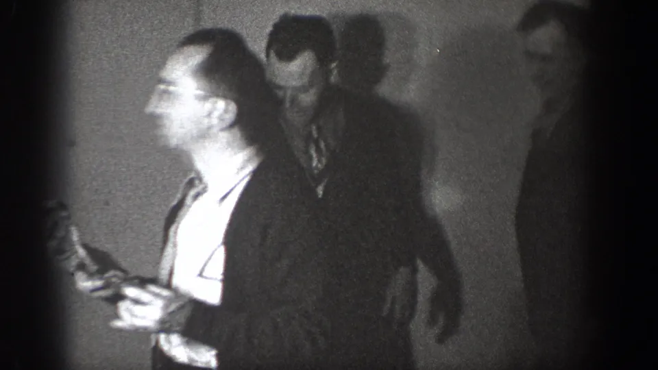 Amateur 8mm circa 1939, Chelan, Washington — man in glasses and tie tilts head upward; second man stands in background against a plain low-lit interior wall.