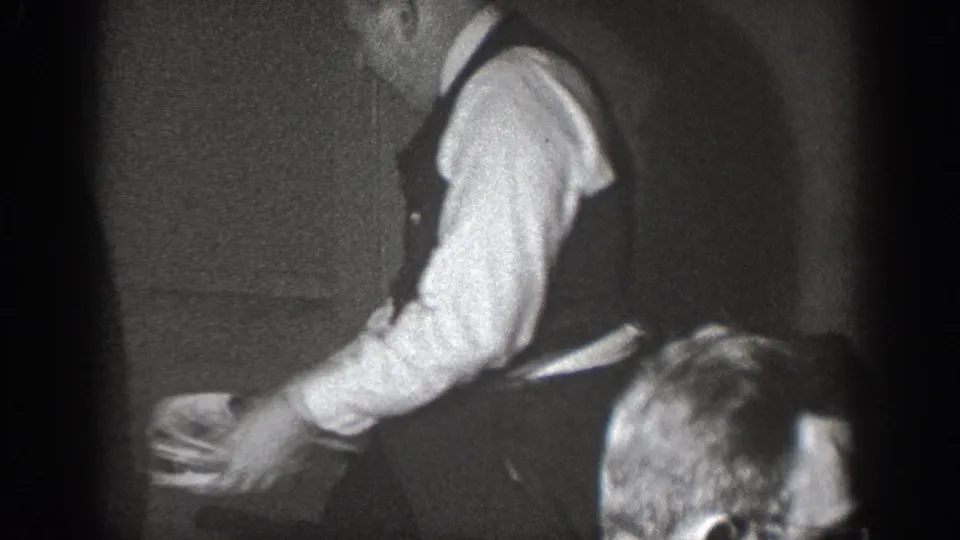 Amateur 8mm circa 1939, Chelan, Washington — man in vest and white shirt bends forward over a dark object; partial figure of a second person appears at the lower frame edge.