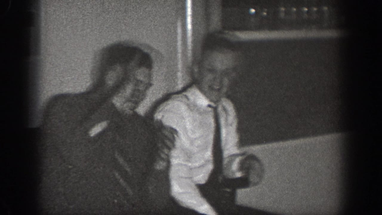 Two men in dark suits and ties laugh together in a domestic interior, Chelan, Washington, circa 1939, filmed on 8mm.